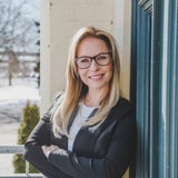 smiling woman with long blonde hair and glasses smiles as she leans against a wall outside of a buliiding